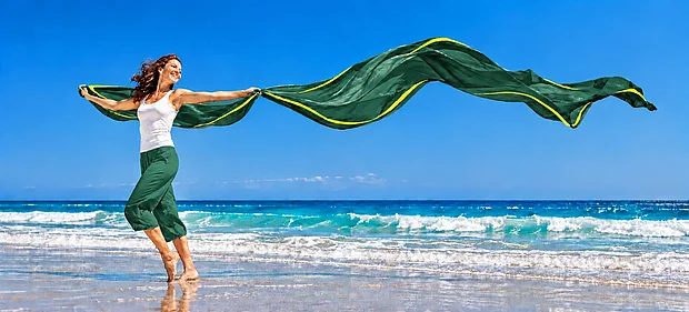 Lächelnde Frau läuft barfuß am Strand und hält ein langes grünes Tuch mit gelben Streifen, das hinter ihr im Wind weht